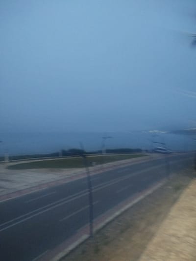 The image presents an outdoor scene at dusk or twilight, overlooking a coastal area in Fnideq, Morocco. A paved road occupies the foreground and midground, featuring multiple lanes marked by white lines. It is bordered by a distinct red-toned curb and green grassy verges. Several dark, slender poles, likely streetlights, are scattered along the roadside, appearing blurred due to motion or focus issues.
Beyond the road, a vast expanse of dark blue water, presumably the sea or an ocean bay, stretches towards the horizon, blending with the uniformly dark blue sky. Faint, scattered lights are visible in the far distance across the water, suggesting a populated area or a city on the opposing shore. A heavily blurred object, possibly a boat or a static structure, is vaguely discernible on the water's surface.
The overall lighting indicates low light conditions, characteristic of twilight or early night. The entire photograph exhibits a significant degree of blur and softness, suggesting it was taken from a moving vehicle or with camera shake. No people or specific activities are clearly visible. The scene conveys a quiet, indistinct evening atmosphere. There is no visible text in the image.