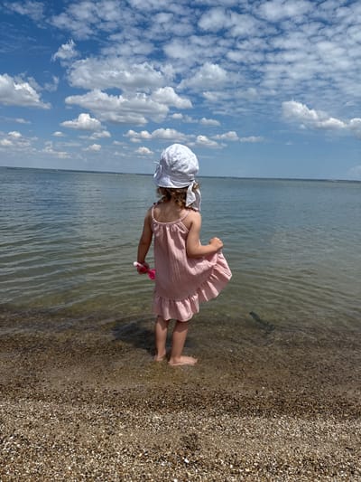 A young child stands at the edge of a body of water, facing away from the camera. The child is wearing a white sun hat and a light pink, ruffled dress. They are holding a pink toy in their right hand. The water is clear and shallow, with a sandy and pebbly shoreline. In the background, the vast expanse of the water meets a hazy horizon under a blue sky filled with fluffy white clouds. The scene appears to be a calm, sunny day, likely at a lake or a calm sea. The location is identified as Sukholuzhzhia, Ukraine.