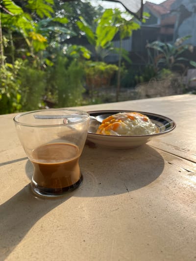 A glass of coffee and a bowl of fried eggs are on a table. The table is made of stone and is located outdoors. In the background, there are trees and houses. The sun is shining, creating a shadow from the glass on the table. The scene appears to be a breakfast setting.