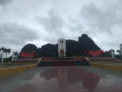 The image captures a grand public square or memorial site in Hong Gai Ward, Vietnam, under an overcast sky. The central feature is a tall, white, modern monument with a golden sphere at its core, flanked by imposing, tree-covered karstic mountains in the background. The sky is filled with grey, heavy clouds, suggesting a cloudy day or recent rain, as evidenced by the wet, reflective tiled ground in the foreground.
Leading up to the monument is a wide, multi-tiered staircase made of dark red and grey steps, reflecting on the polished surface below. On either side of the stairs, at a lower level, are manicured flower beds bursting with purple blooms, interspersed with large, stylized red structures resembling open lotus flowers or flags, creating a vibrant contrast against the muted tones of the sky and mountains. Beyond these, palm trees are visible, adding to the tropical setting.
The foreground consists of a large, expansive area of wet, reflective pavement, mirroring the sky, the monument, and the surrounding structures. On the left side of this paved area, near a yellow retaining wall, some cleaning equipment, possibly a mop or squeegee and a red bucket, is casually placed. No people are visible, lending a sense of quiet grandeur to the scene. The overall atmosphere is one of serene solemnity, with the majestic natural landscape providing a dramatic backdrop to the contemporary monument. No discernible text is visible in the image.