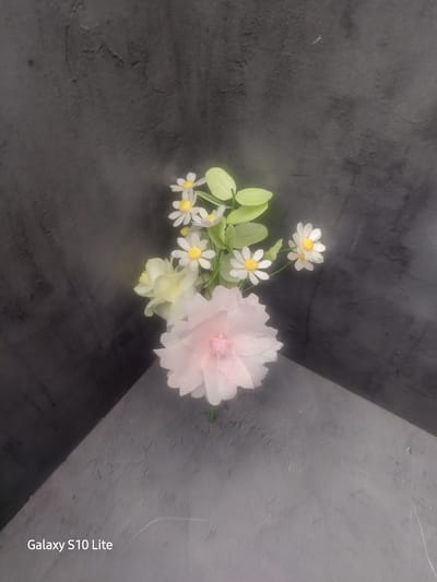 A close-up, top-down shot features a small arrangement of artificial flowers against a dark, textured background and a grey surface. The bouquet includes several small white daisy-like flowers with yellow centers, some light green leaves, and a larger, pale pink layered flower. The flowers are positioned on the right side of the frame, with the pink flower slightly lower and to the left of the smaller daisies. The background appears to be a dark grey wall with a rough texture, possibly concrete or stucco, and the foreground is a grey surface that transitions from dark to light, suggesting a table or platform with lighting that casts a shadow from the bouquet. A watermark "Galaxy S10 Lite" is visible in the bottom left corner. There is no discernible location in Wellingborough, United Kingdom, or any context suggesting this specific place. The image is a still life composition of artificial floral elements.