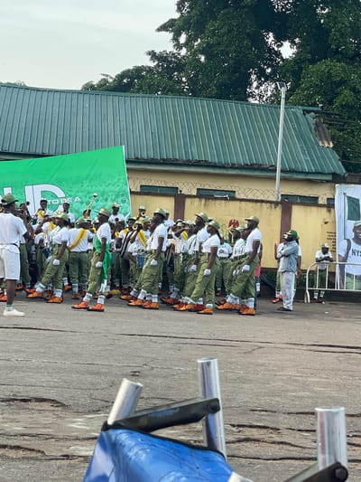 A group of people, likely young adults, are lined up outdoors in what appears to be a parade or formal assembly. They are dressed in matching uniforms consisting of olive green trousers, white shirts with green trim, and green caps. Many are wearing orange boots. Some individuals are holding musical instruments, suggesting a band or marching ensemble.
The scene is set in an outdoor paved area with a building featuring a green corrugated metal roof in the background. A large green banner with white lettering is prominently displayed. The sky appears overcast.
Visible text on the banner includes "TO BE A" and partial words like "Youths ab" and "Let us lif". Another banner in the background, partially obscured, shows "NYS" and "Nigeria". The overall atmosphere seems organized and possibly ceremonial. The time of day appears to be daytime, given the natural light. The location is identified as Alagba, Nigeria.