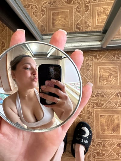A young woman is taking a selfie in a small circular mirror that she is holding in her left hand. The mirror reflects her face, which is contorted into a pout, her right arm, and the top of her chest. She is wearing a white tank top. She has a ring on her left ring finger. Her nails are long and polished. Behind her, on the floor, is a black slipper with a crescent moon design and the words "MEET ME" embroidered on it. The floor appears to be tiled with a brown pattern. A metal frame, possibly a window or door frame, is visible to the left of the frame, with some light reflecting off of it. The overall scene suggests a casual indoor setting. There is no indication of time of day or weather.