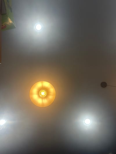 The image shows a ceiling with several lights. In the top left corner, a portion of a green box with yellow lettering is visible. There are three visible light sources on the ceiling: a bright white circular light in the upper middle, a warm yellow circular light with visible filament in the middle, and another bright white circular light in the lower right. A small dark circular object is visible in the far right of the image, attached to the ceiling by a thin wire. The overall scene is indoors, illuminated by artificial light. There are no people or animals present. There is no indication that this image is from Шебунино, Russia.