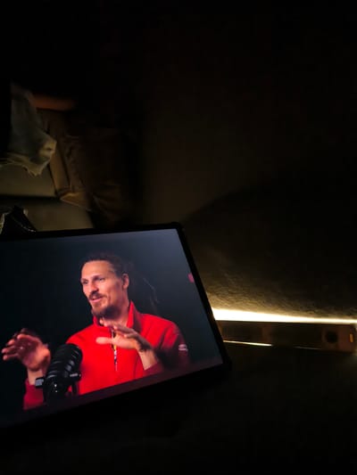 The image shows a tablet screen displaying a man with dreadlocks wearing a red zip-up shirt speaking into a microphone. His hands are raised in a gesturing motion. The tablet is placed on a dark surface, likely a table or desk. To the right of the tablet, a bright, horizontal light source illuminates the dark background. Above the tablet, a blurry figure is partially visible, lying down and covered with a light-colored cloth. The scene appears to be in a dimly lit room. The location is specified as Pendik, Turkey.