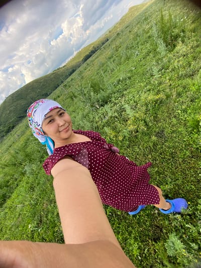 The image shows a young woman lying on a grassy hillside in Maloarslangulovo, Russia. She is wearing a maroon polka dot dress, a white headscarf with colorful patterns, and blue slippers. The woman is smiling and looking at the camera, suggesting a casual and happy mood.

The setting is a lush green landscape with rolling hills stretching into the distance. The sky is partly cloudy, with bright white clouds against a blue sky, indicating it is likely daytime. The sun appears to be shining, casting soft light on the scene. The woman's pose and the camera angle suggest she is taking a selfie. The grass is green and appears to be growing in a natural, uncultivated area, possibly a meadow or pasture. There are small yellow wildflowers scattered throughout the grass.

The overall impression is one of a peaceful and serene moment in nature. The woman seems relaxed and content, enjoying the outdoors. The image captures a simple yet beautiful scene of a person connecting with the natural environment. No text is visible in the image.