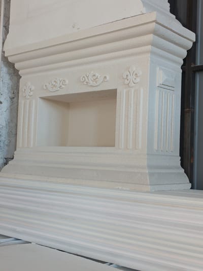 This media file is a medium-shot, eye-level photograph of a white, decorative architectural element, likely a fireplace surround or a pedestal, made of stone or a similar material. The object is positioned in the foreground, dominating the frame, and appears to be unfinished or in a workshop setting, as suggested by the presence of other materials in the foreground that resemble stacked molding or trim pieces.

The architectural element features intricate carvings, including fluted columns on its sides, decorative scrollwork above a central recessed niche, and a stepped, ornate base. The overall design suggests a classical or baroque style. The texture of the material is visible, appearing somewhat rough or porous.

In the background, to the right, a dark metal grate or window frame can be seen, hinting at an indoor or semi-covered location. To the left, a distressed, textured wall, possibly brick or plaster, is partially visible, showing signs of wear and tear. The lighting is even, suggesting an overcast day or indoor illumination, with no strong shadows. The color palette is predominantly white and beige, with some muted grays and browns in the background. There is no discernible activity or event taking place; the focus is solely on the static object. No people are visible in the frame, and there is no visible text. The location context provided is Durlești, Moldova.