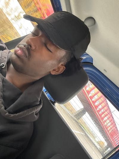 An image, tilted clockwise, depicts a male individual with dark skin asleep or resting inside a vehicle in what is identified as Dettwiller, France. The man's head is tilted to his right, supported by a black headrest, his eyes closed. He wears a black baseball cap with a silver-colored trim and a dark grey hooded sweatshirt. A mustache and short beard are visible. The vehicle's interior features a light-colored ceiling with a circular light fixture and blue trim along the top edge of the rear window. Through the rear window, a blurred outdoor scene is visible, including what appears to be a road or paved area with distant foliage on the left. On the right, a large red object with indistinct white markings is partially visible, suggesting a commercial structure or a large vehicle in the external environment of Dettwiller, France.