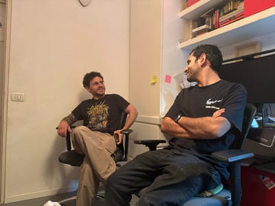 An indoor scene in Ramat HaSharon, Israel, depicts two adult males seated in office chairs, appearing to engage in conversation or observe something to their right. The male on the left, with curly brown hair, a short beard, and glasses, smiles while looking to his right. He wears a black t-shirt with a multi-colored graphic design, light brown cargo pants, and has tattoos on his left forearm. His legs are crossed, and his left hand rests on his leg. The male on the right, with short dark hair, smiles while looking to his left. He wears a black t-shirt with a white graphic, dark pants, and a dark smartwatch on his left wrist. His arms are crossed over his chest, and a green cushion is visible on his chair. The background features a plain light-colored wall with a light switch on the left. To the right, behind the second male, a white shelving unit filled with books and red containers is visible. A dark computer monitor, a "Scorpion" branded mousepad, and other office equipment are on a desk. A yellow and a pink sticky note are adhered to a nearby wall or cabinet. A red object is on the floor or a lower shelf.