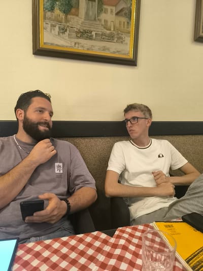 Two men are seated at a table covered with a red and white checkered tablecloth. The man on the left has dark hair and a beard, and is wearing a grey t-shirt with a logo. He is holding a black smartphone in his left hand and has a beaded bracelet on his right wrist. The man on the right has blonde hair and is wearing glasses and a white t-shirt with a logo. He is looking to his left, with his hands clasped. Behind them, a dark brown upholstered bench is visible, and a framed painting hangs on the wall above. The setting appears to be a cafe or restaurant in Cetinje, Montenegro.