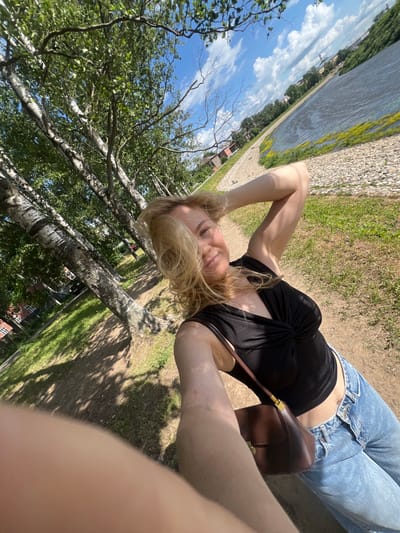 A young woman with blonde hair takes a selfie outdoors on a sunny day. She is wearing a black top and blue jeans, and is carrying a brown purse. She is standing near a path with trees on her left and a body of water on her right. The location appears to be a park or waterfront area in the city of Vologda, Russia. The trees are likely birch trees, characterized by their white bark with dark markings. The water body is likely a river or a lake, with a gravelly bank lined with green grass and some yellow wildflowers. In the background, there are buildings and more trees, suggesting an urban or semi-urban environment. The sky is blue with scattered white clouds.