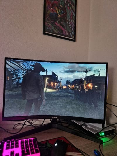 The image shows a computer monitor displaying a video game. The scene in the video game is an evening shot of a town with a dirt road and wooden buildings on either side. A lone figure in a black coat and hat is standing in the middle of the road. A black computer mouse is lying next to a keyboard on a desk in the foreground. The monitor is on a stand and is connected to wires that run to a small black box and a green light. The image was taken in a bedroom in Kernen im Remstal, Germany.