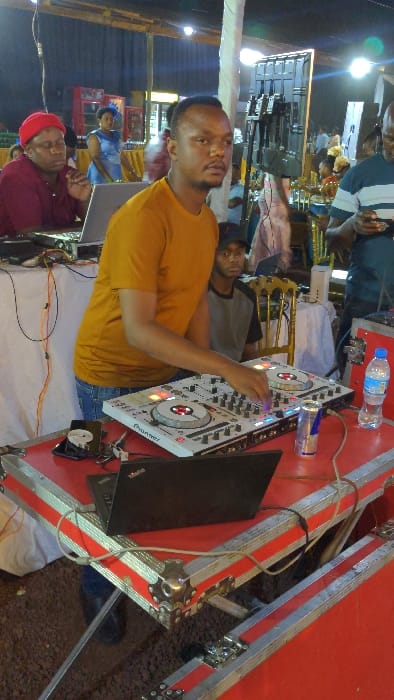 A man in an orange shirt operates a Pioneer DJ mixer on a red and silver stage platform. A laptop sits adjacent to the mixer. Behind the DJ, a crowd is visible, and another man looks on. A water bottle and Red Bull can are near the DJ equipment. The image is somewhat blurry, taken at an outdoor event during nighttime.