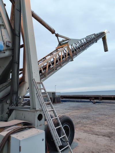 The image depicts a large, industrial conveying system in the city of Shcheleiki, Russia. The system appears to be on a dock or pier, as the water and horizon are visible in the background. The main components are a large, gray metal structure with a ladder attached, a conveyor belt extending upwards at an angle, and a wheel and control panel near the base. The sky is overcast, suggesting a cloudy day. There are no people visible in the image.