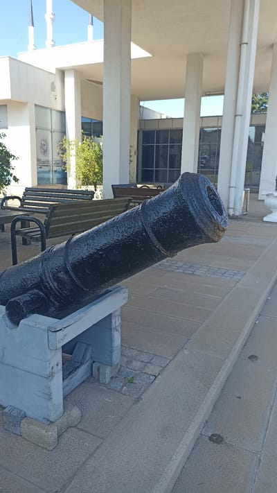 The image shows a black, antique cannon positioned on a light blue wooden stand. The cannon is the primary subject, with its barrel extending diagonally from the lower left to the upper right of the frame. The barrel has a textured, glossy finish and appears to be made of metal. It is mounted on a wooden carriage that is supported by concrete blocks.
In the background, there is a modern building with large windows and white columns. The architecture suggests an outdoor public space. Several wooden benches are visible in front of the building, implying a seating area for visitors. Trees and greenery are present between the benches and the building. The sky is clear and blue, indicating a bright, sunny day. The paving stones in the foreground and midground suggest an outdoor plaza or walkway. The overall setting appears to be a historical site or a public park in a city, possibly near the coast given the imagery seen through the windows, depicting ships. There are no people visible in the frame, and no text is discernible. The time of day appears to be midday, given the bright sunlight and the shadows.