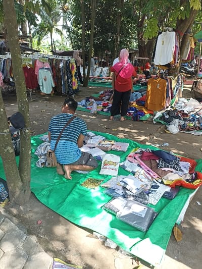 This outdoor market scene in Mataram, Indonesia, captures people browsing and selling various clothing items under the dappled shade of trees. In the foreground, a man sits barefoot on a green tarp, facing away from the camera. He wears a blue and white striped t-shirt and dark shorts, with a brown shoulder bag slung across him, and appears to be arranging or inspecting folded clothes displayed on the tarp. Among the items on his tarp, a white bag with "FASHION" and "COLLECTION" printed on it, along with a tropical graphic, is visible, as well as a packaged item partially showing "SPRING SUMMER".
To his right, in the mid-ground, a woman stands with her back to the viewer, wearing a bright red top, dark pants, and a pink head covering or shawl. She is observing clothing piles and items on another green tarp. In the background, more clothing racks display garments, and other shoppers or vendors can be seen. Some clothing racks feature price tags, with visible text indicating "RP. 35.000" and "RP. 45.000". The market is set on a dirt ground with some paved areas, suggesting an informal selling environment. The bright sunlight filtering through the trees indicates a clear, sunny day, likely morning or early afternoon.