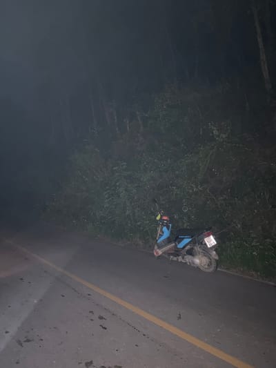 This image, taken in Ban Mae Ping, Thailand, captures a nighttime scene on a paved road bordered by dense vegetation. A single blue and black scooter, marked "SEMPEX" and bearing a Thai license plate displaying "9418", is parked on the dirt shoulder to the right of the road. The scooter exhibits dirt on its chassis and wheels, and an unknown item, possibly gear or luggage, is secured to its front. The asphalt road features a continuous yellow line near its center and shows dark, irregular stains. To the right of the scooter and extending into the background, a thick growth of bushes and trees is visible, with distant trees partially obscured by an atmospheric haze or fog. No individuals are present in the frame. The overall lighting is dim, indicative of darkness.