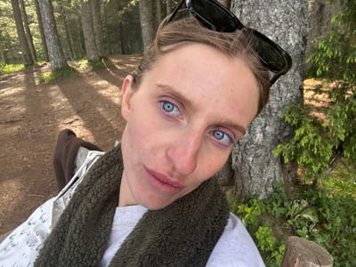 A young woman is pictured in a close-up selfie, her face is framed by a thick, dark green, fuzzy scarf or vest. She is wearing a light gray sweatshirt. Her hair is pulled back, and she has black sunglasses resting on top of her head. She has bright blue eyes with purple eyeliner. Her skin appears fair with some redness and freckles. Her lips are closed and slightly parted.
The background suggests a natural, outdoor setting. Tall, slender trees with textured bark dominate the scene, with dappled sunlight filtering through their branches, casting shadows on a dirt path. The ground is covered with pine needles and dry leaves, indicating a forest or wooded area. Green foliage, including ferns and smaller plants, grows around the base of the trees and in the foreground. A weathered wooden post is visible on the right side of the image. The overall impression is of a serene, wooded environment. The specific location in Pitomine, Montenegro cannot be determined from the visual information alone.