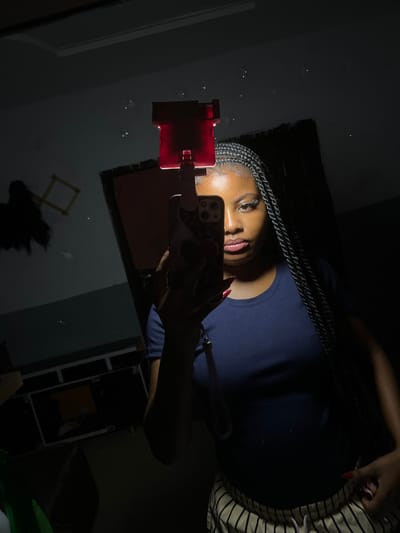 A forensic examination of the image reveals a selfie taken in a mirror within a dimly lit indoor environment in Ipetumodu, Nigeria. The primary subject is a dark-skinned female, partially illuminated by a clip-on light attached to her smartphone. She is depicted from the upper chest to just below the waist, facing slightly to the viewer's right. Her hair is styled in long, dark braids. She wears a dark blue short-sleeved t-shirt and light-colored pants with thin black vertical stripes and a white drawstring. Her fingernails are painted red. Her left hand holds a dark-colored smartphone, equipped with a red-tinted clip-on light that projects illumination onto her upper face, including her eyes and lips which show cosmetic enhancement. Her right hand is visible near her right hip, touching her pants. The background, visible through the mirror's reflection, includes a dark wall, scattered bright speckles in the upper left, an angular light-colored frame structure, and a dark rectangular object with horizontal lines at the bottom left, possibly a case or storage unit.