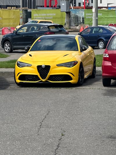 The image shows a yellow Alfa Romeo car parked on an asphalt surface in what appears to be a parking lot. The car is the primary subject, positioned in the foreground and facing towards the viewer. Its front grille, headlights, and hood are clearly visible. In the background, other vehicles are present, including a black SUV on the left, a blue sedan on the right, and parts of other cars are visible. Yellow and green banners with Cyrillic text are displayed behind the vehicles. The environment suggests an outdoor urban or suburban setting. The specific location is identified as Бараўлянскі сельскі Савет, Belarus, implying this is a public parking area within that administrative division. There are no people visible in the image.