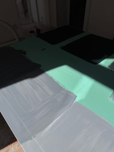 The image shows a close-up of a surface, possibly a wall or board, with different colors painted on it. There are rectangular sections painted in black, light green, and grey. The paint application appears uneven, with visible brushstrokes in the grey sections. A diagonal shadow cuts across the surface, adding contrast. The image includes a corner of a room with a white door and some construction elements visible in the background. The information provided does not allow me to determine the precise location in the city of Erba, Italy.