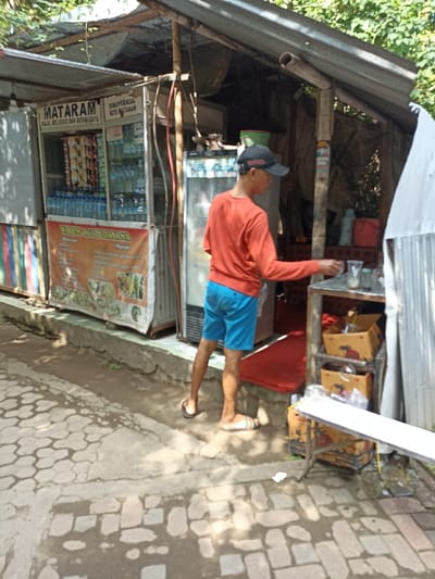 This media file captures an outdoor scene featuring a small street vendor stall, likely a "warung," in Mataram, Indonesia. The stall, constructed from corrugated metal sheets and wooden supports, is situated on a raised concrete platform next to a paved pathway with a cobblestone-like pattern.
In the center, a person, possibly a vendor, is seen from a rear-side angle. He wears an orange long-sleeved shirt, blue shorts, and flip-flops, topped with a black baseball cap. He is standing at a small, rustic wooden counter inside the stall, reaching towards some glasses, suggesting he is preparing a drink or attending to customers.
To the left of the person is a glass display case or refrigerator filled with numerous bottles of water and various snack packets. Below this display, a banner is prominently displayed, reading "WARUNG A ZIDNI MANA" and "Makanan" (Food), accompanied by images of different food dishes. Above the display, text includes "MATARAM," "MAJU RELIGIUS DAN BERBUDAYA," and "DISPERINDAG KOTA MATARAM," identifying the city and possibly a local government endorsement. To the person's right, another, taller refrigerator is visible, along with shelves holding cardboard boxes and other items. A red mat is placed on the ground inside the stall entrance.
The scene is brightly lit by natural sunlight, casting shadows on the ground, indicating daytime. Green foliage from trees is visible in the background, suggesting an outdoor, possibly semi-urban or residential setting. The overall impression is that of a typical local eatery or convenience stall.