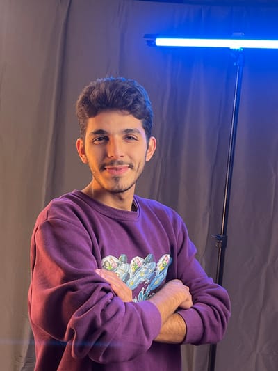 A young man smiles, arms crossed, wearing a purple sweater with a light blue and white graphic. He has brown hair and a light beard. The background consists of a dark gray sheet and a long light emitting blue light. The man is positioned slightly off-center.