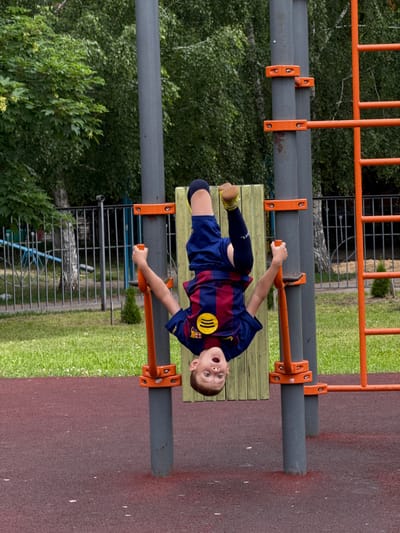 A young boy, wearing a blue and burgundy soccer jersey with yellow accents and matching shorts, is depicted in a playful pose on an outdoor playground. He is upside down, hanging from a horizontal bar with one arm extended and the other bent. His mouth is open in what appears to be a shout or gasp of excitement, and his eyes are wide open, looking towards the viewer. The boy is wearing dark blue socks and yellow shoes.

The playground equipment is made of metal, with orange supports and grey horizontal bars. A wooden bench is visible below him, also attached to the metal structure. The background consists of lush green trees and grass, suggesting a park or recreational area. In the far background, a metal fence and some darker structures are visible. The ground beneath the playground area is a reddish-brown, shock-absorbent surface, common in modern playgrounds.

The lighting suggests it is daytime, likely a sunny or overcast day given the even illumination. The overall scene conveys a sense of fun and energetic play. The boy's expression and posture indicate he is actively engaged in a game or activity on the playground. The location cues, such as the type of playground equipment and the surrounding vegetation, are consistent with a park or a schoolyard. While the specific location is Stary Oskol, Russia, this cannot be visually deduced from the image itself. No visible text is present on the boy's clothing or the playground equipment.
