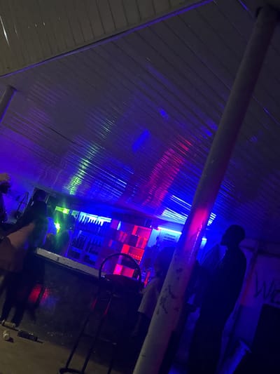 The image depicts an indoor bar scene illuminated by vibrant blue and pink neon lights, casting colorful reflections on the corrugated metal ceiling and surrounding surfaces. Several individuals are present, though their features are obscured by the low lighting and silhouettes. In the background, shelves stocked with bottles are visible behind a counter, suggesting it is a functional bar. A stool is positioned near the counter. Graffiti-like text, including "WE", is present on a wall to the right. The overall atmosphere suggests a nighttime setting within a bar or lounge, possibly located in Wumat, Nigeria, given the context provided. The scene is dimly lit, with the neon lights creating a dynamic and somewhat abstract visual effect.