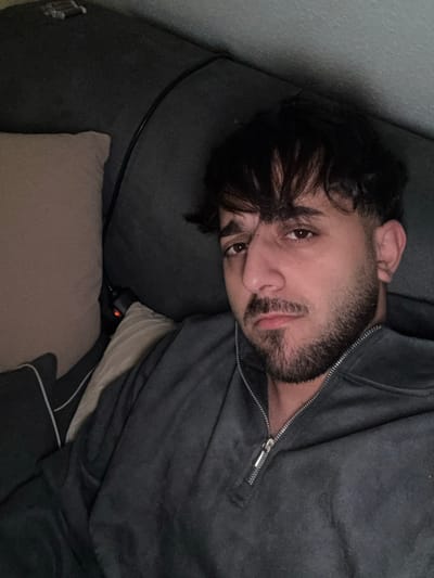 A person lies on a dark gray couch, head resting on a matching cushion. The individual has dark hair with bangs, a dark beard, and brown eyes, and is wearing a gray zip-up sweater. A tan pillow is also visible on the couch. A black cord stretches behind the person's head along the couch. Part of a bed is beneath the person on the couch. The scene has a muted, dimly lit atmosphere.