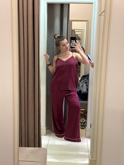 In a fitting room, a young woman stands in front of a large mirror, wearing maroon silk pajamas. She is holding a phone in her right hand, seemingly taking a selfie, and has her left arm bent at the elbow with her hand raised in a peace sign. Her hair is pulled up in a bun, adorned with a scrunchie. She is wearing a necklace and bracelets. In the mirror's reflection, a series of repeating doorways and mirrors create an optical illusion. A brown curtain hangs on the left side of the frame, and a beige upholstered stool is visible in the foreground. The room appears to be in a clothing store or boutique. The location is not specified in the image or hints, therefore it is not possible to determine if it is in Vladimir, Russia.