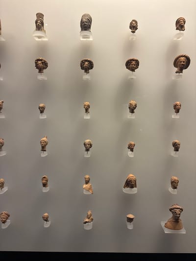 This media file displays a grid of ancient terracotta busts, likely exhibited in a museum in Istanbul, Turkey. The busts are small, predominantly featuring human heads with varying hairstyles and facial features, suggesting they might be representations of individuals or deities from antiquity. They are mounted on clear, minimalist stands against a plain, light-colored wall, which serves as a neutral background to emphasize the artifacts.
The lighting is even and diffuse, illuminating the details of each bust without casting harsh shadows, and the overall presentation creates a sense of order and historical significance. The objects are arranged in neat rows and columns, with some rows having more busts than others, creating an irregular but intentional pattern. Some busts are positioned at slight angles, and their varying sizes and details imply a range of origins or periods. The style of the busts, characterized by their material and depiction of faces, points to a classical or Hellenistic influence. No text is visible in the image. The context of Istanbul, a city with a rich historical tapestry that bridges Eastern and Western civilizations, makes this display a compelling glimpse into ancient art and culture.