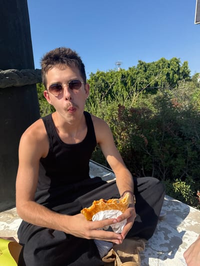 A young male is seated outdoors in bright daylight, assumed to be in Kfar Vitkin, Israel. He wears a black ribbed tank top, loose-fitting black pants, and oval sunglasses with dark lenses. His short, dark hair is visible, and he has a thin mustache. He holds a partially eaten golden-brown pastry with a reddish filling, wrapped in white paper, with both hands. His mouth is puckered forward, and a small amount of food residue is visible on his lower lip. A dark, cylindrical structure with a rope wrapped around it is visible on the left. A yellow bag is partially visible on the ground to the lower left, and a brown paper bag with a clear plastic cup is visible to the lower right. The background features dense green trees and bushes under a clear blue sky, with a distant communications tower visible through the foliage.