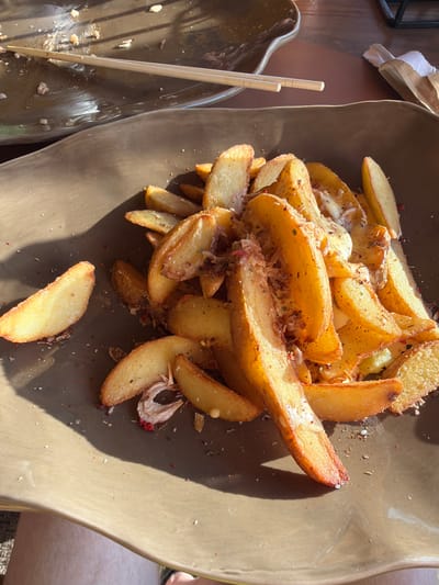A top-down, medium shot shows a large, flat, brown ceramic plate filled with golden-brown potato wedges, seasoned with herbs and spices. The wedges are irregular in shape, indicating they are likely hand-cut. Some wedges appear to be topped with a creamy white sauce or cheese. A few scattered pieces of what appear to be dried garlic or shallots are visible among the potatoes. The plate is on a wooden surface, suggesting an outdoor or rustic setting. To the left of the main plate, a portion of another brown ceramic dish is visible, containing two wooden chopsticks and remnants of food. The lighting suggests it is daytime, with strong shadows cast by the potato wedges, indicating sunlight. The overall impression is of a casual meal, possibly at a restaurant or an outdoor gathering. No visible text is present.