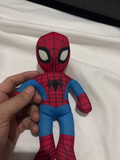 A plush Spider-Man toy lies on a white, slightly wrinkled surface, possibly a bed sheet. The toy's head is red with web patterns and large white eyes. The body is red and blue, also featuring a spider web design and a black spider emblem on the chest. A human hand partially obscures the toy's midsection. The image is taken from a slightly elevated angle. A dark-colored object is vaguely visible in the upper left corner of the frame.