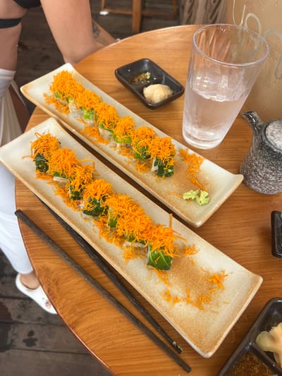 Two rectangular plates of sushi rolls are presented on a wooden table at a restaurant in Ratsada, Thailand. The sushi rolls are topped with shredded orange vegetables, resembling crispy fried carrots or sweet potato strings. Each plate also has a small dollop of green wasabi paste. In the background, a glass of water with ice is visible, along with a small grey and white teapot and a black dipping sauce dish containing soy sauce. Chopsticks are laid across the plates. A person's legs and feet are visible on the left side of the image, with white pants and footwear. The table surface has a distinct wood grain pattern. The flooring appears to be a combination of wood and possibly concrete.