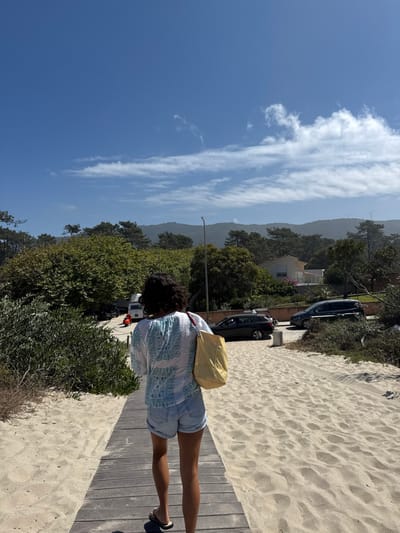 The image captures a female subject walking away from the camera on a weathered wooden boardwalk that traverses a sandy area in Caminha, Portugal. The subject wears a white, long-sleeved top with light blue patterned embroidery on the back, light blue denim shorts, and dark flip-flops, carrying a yellow and white striped tote bag on her left shoulder. The boardwalk is bordered by light-colored sand exhibiting footprints and sparse green and brown vegetation. In the midground, a paved road is visible beyond the sandy area and a line of green trees. Several vehicles are present on the road, including a dark station wagon, a dark SUV, a white van, and a red moped. A street light pole stands near the road. Further in the background, a white house with an orange roof is partially visible amidst more green trees. Distant green mountains rise under a clear blue sky, featuring scattered white clouds and two white wind turbines visible on the far right peak. The scene occurs under bright daylight conditions.
