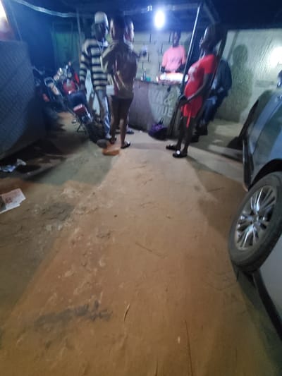 The image shows an outdoor scene in Nnewi, Nigeria, at night. The area is illuminated by a light source in the upper left of the image. A car is parked at an angle in the upper right of the image. There are at least two people visible. One individual wears a striped shirt and stands beside a motorcycle. Another person wears a red shirt and stands near the top of the image. The ground is a mix of dirt and concrete.