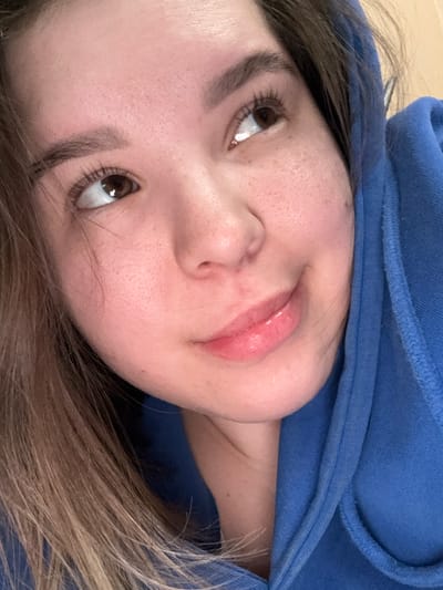 A close-up portrait features a young adult female. Her skin is fair, exhibiting visible pores and sparse freckling on the cheeks and forehead. She has brown eyes, naturally shaped eyebrows, and pronounced eyelashes. Her lips are full with a light, glossy application, curving into a subtle, closed-mouth smile. A small blemish is located on her right cheek. Light brown hair with blonde highlights is visible on the left side of the frame. She is clad in a blue hooded garment, likely a hoodie, with the hood partially obscuring the right side of her head and neck. Her gaze is directed upwards and to the right. The background is uniformly light-toned and out of focus, indicating an indoor environment. This scene is situated in Nizhnekamsk, Russia. No specific visual elements within the image confirm this precise geographic location.