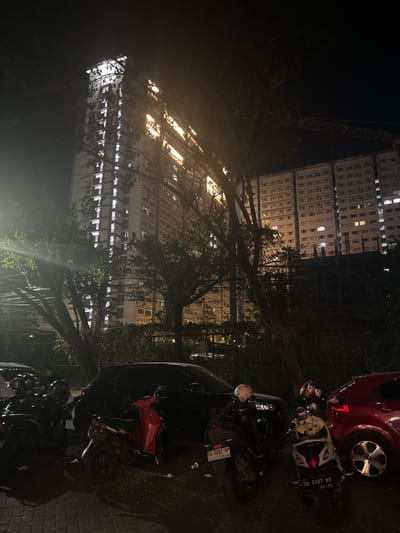 In the city of Makassar, Indonesia, at night, a tall, brightly lit apartment building stands in the background. In front of the building, several motorcycles are parked. Some of the motorcycles are black, one is red, and another is white and black. There are also two cars, one black and one red, parked next to the motorcycles. Trees with dark leaves obscure parts of the buildings. A construction crane is visible in the upper right corner of the image. The ground is paved with cobblestones.
