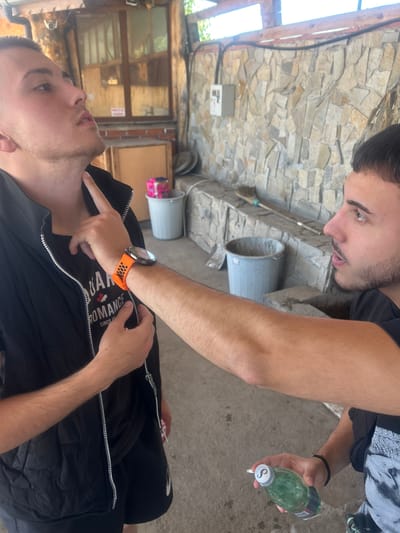Two men are outdoors in what appears to be a courtyard or patio area. The location is Elhovo, Bulgaria. One man is lying on the ground wearing a black t-shirt and a black vest. He has a smartwatch with an orange band on his left wrist. The other man is standing, looking down at the man on the ground. He is holding a green bottle in his right hand. The courtyard has stone walls, a concrete floor, and several buckets are present. There is an electrical box on the wall. The scene suggests a moment of concern or assistance.