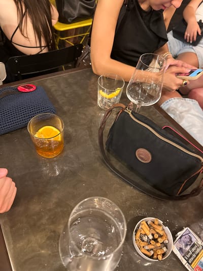 A medium shot captures a table with various items, including drinks, bags, and a ashtray filled with cigarette butts, suggesting a casual gathering. The scene appears to be indoors, possibly a bar or restaurant. A woman in the background is seen holding a smartphone and wearing jewelry. In the foreground, a glass with ice and an orange slice is visible, along with a dark blue knitted bag. A black clutch bag with a leather strap and a circular logo rests on the table. The floor or table surface is dark and shows signs of wear. A pack of cigarettes with Italian warnings is also present. The environment suggests a social setting where drinks and cigarettes are being consumed. The lighting is dim, typical of an evening or indoor establishment. The overall impression is one of relaxed social interaction.