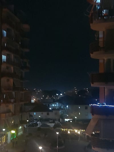The image captures a nighttime urban scene, likely from an elevated perspective overlooking a street or waterfront. In the immediate foreground, several rows of empty chairs or seating arrangements, possibly belonging to an outdoor restaurant or public viewing area, stretch across the lower portion of the frame. Beyond these structures, a dark expanse, consistent with a body of water or a very dark sky, occupies the upper-left area of the rotated image.
To the right, a street or promenade runs parallel to this dark expanse, lined with buildings of various sizes. These buildings display illuminated windows, and streetlights cast pools of light on the thoroughfare. Faint outlines of vehicles can be seen on the street. Decorative string lights contribute to the nighttime ambiance; notable are blue string lights draped along a structure on the left side, and yellow/orange string lights illuminating what appear to be trees or bushes on the right.
The scene is devoid of clearly identifiable people or specific activity, conveying a quiet, peaceful atmosphere typical of a city after dark. The lighting is entirely artificial, highlighting architectural features and adding a festive touch with the string lights. No discernible text is visible. Given the context, this could be a view of a lakefront or riverfront area in Tiranë, Albania, such as the Artificial Lake of Tirana, at night.