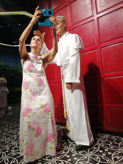 A woman in a floral sundress is taking a selfie with a wax figure of Pope John Paul II. She is holding up a blue tablet to take the picture. The location is in Hong Gai Ward, Vietnam. The background includes a red wall and a patterned black and white floor. There is a person in the background to the left.