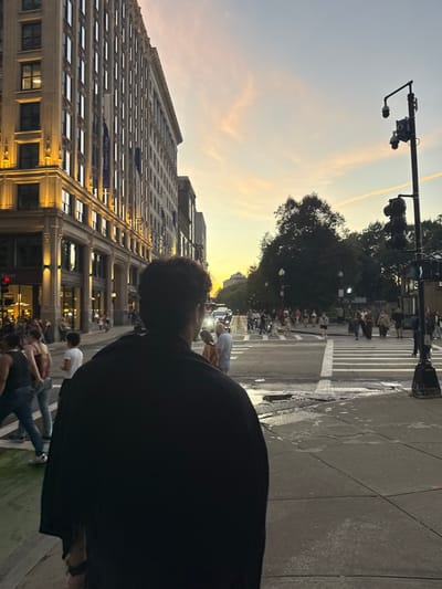The image depicts a dusk scene at a city intersection in Boston, Massachusetts, United States. In the foreground, a person with dark, curly hair, viewed from behind, stands on a concrete sidewalk wearing a dark, possibly black, garment. To the left, a tall, multi-story building with "Emerson College" banners displayed vertically on its facade is brightly lit on its lower levels. The street in the midground features multiple lanes, white crosswalk markings, and several puddles on the asphalt surface. A green-painted bike lane is visible on the far left. Numerous pedestrians are either crossing the street, waiting on sidewalks, or congregated near the Emerson College building. Several vehicles with illuminated headlights are present on the street. On the right, a tall black pole supports traffic lights and at least two surveillance cameras. Beyond the street, a dense cluster of trees signifies a park area, likely Boston Common. The sky exhibits sunset colors, transitioning from orange and pink near the horizon to light blue above, with faint clouds and a distinct contrail.