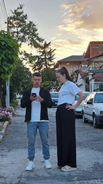 In a residential street in Karlovo, Bulgaria, two individuals stand on a paved surface under a late afternoon or early evening sky with golden-hued clouds.
On the left, a male in his 20s, wearing a white t-shirt, black zip-up jacket, light blue distressed jeans, and white sneakers, holds a black smartphone in both hands, looking intently at its screen. A watch is visible on his left wrist. His mouth is slightly agape.
To his right, a female in her 20s, with dark hair pulled into a high ponytail, wears a white t-shirt, black wide-leg trousers, and light-colored platform sandals. Her right hand rests on her hip, and her left hand is similarly positioned. She looks downwards and slightly to her left, towards the male or the ground. A silver bracelet is on her left wrist.
The foreground consists of a rough, paved street surface. To the left, a low concrete curb borders a strip of soil with pink flowering plants and dense green foliage covering a vertical structure. In the background to the left, green-leaved trees are visible.
To the right, parked vehicles line the street, including a dark-colored car with a Bulgarian license plate (PB 1XXX) and a white older model sedan. Behind the vehicles are residential buildings with various facades, including exposed brick and white plaster, and some with red-tiled roofs. Overhead power lines traverse the upper portion of the scene, with several trees, including conifers, silhouetted against the bright sky.