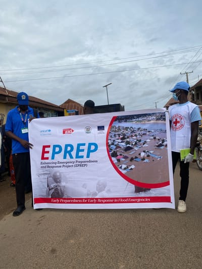 In Rigachikun, Nigeria, two men stand holding a large banner that reads "EPREP: Enhancing Emergency Preparedness and Response Project." The banner also features logos from UNICEF, Christian Aid, SEMA, and the European Union. The image on the banner depicts a flooded town with houses submerged in water. One man on the left wears a blue polo shirt with an ID card and a blue cap. The man on the right wears a white vest with the Nigerian Red Cross logo, a light blue t-shirt, a blue cap, a face mask, and gloves. They are standing on a paved street with buildings and overhead wires in the background. The sky is overcast.