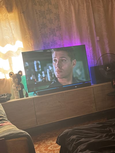 The image captures an indoor scene, likely a living room, at night. A large television screen displays a close-up of a man's face, possibly from a movie or TV show. The room is illuminated by the glow of the TV and a purple light source behind it, casting a colored hue on the patterned curtains. To the left of the TV, on a wooden entertainment stand, are decorative items including a bear-shaped figurine, a small bottle with reeds, and a toy car. A black soundbar sits below the TV. To the right, a black oscillating fan is positioned next to a black box, possibly a speaker or router, which is partially obscured by the fan. The overall impression is a domestic setting, with emphasis on entertainment and ambient lighting. There is no information in the image to determine its location in Makiivka, Ukraine.
