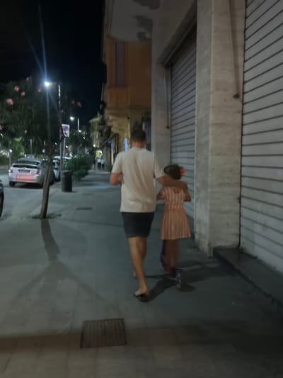 The image displays a nocturnal street scene in Sestri Levante, Italy. Two individuals, an adult male and a child, are observed from behind, walking on a paved sidewalk. The male is wearing a light-colored short-sleeved shirt, dark shorts, and dark flip-flops, with his right arm draped over the child's left shoulder. The child, wearing a light-colored striped dress, is positioned to the male's left and appears to be on a scooter, with two wheels visible near her feet. Her left arm is around the male's waist.
To the right of the subjects, a building facade features a large, closed, horizontally corrugated metal security shutter. Further along the street on the right, other buildings with lighter facades are visible. To the left of the sidewalk, a street with several parked vehicles is present, including a light-colored hatchback. The street is lined with trees bearing sparse foliage and light-colored blossoms, as well as streetlights, a dark cylindrical trash bin, and a traffic sign pole. The scene is lit by artificial streetlights, casting shadows. A rectangular metal grate is visible on the sidewalk in the foreground.