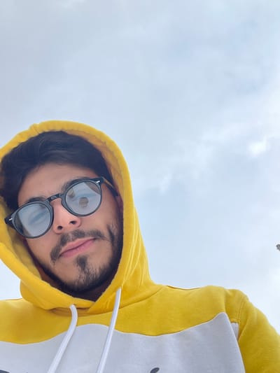 The image depicts a young adult male, positioned at a low angle and looking directly into the camera against an overcast sky. He has dark hair, a beard, and a mustache, and is wearing round-framed black sunglasses. Visible dimples are present on his cheeks. He is dressed in a yellow hooded sweatshirt with a prominent white horizontal stripe across the chest and white drawstrings. The hood is pulled up, framing his head. The background consists predominantly of a light grey, cloudy sky. A faint, indistinct, light-colored structure or object is minimally visible on the far right edge of the frame against the sky. The scene is located in Al-Muhajerin Municipality, Syria.